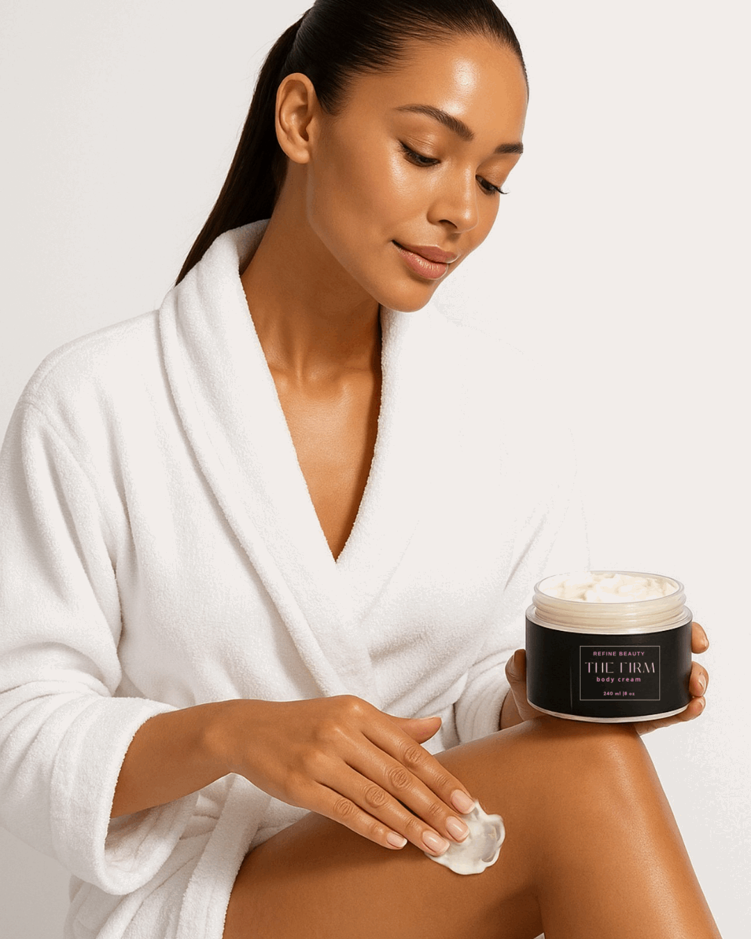 Woman in a white robe applying Refine Beauty cream from a jar labeled 'The Firm body cream on her leg 