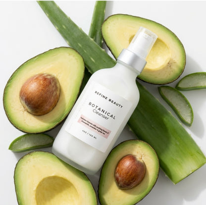 Skincare bottle labeled Refine Beauty Botanical Cleanser surrounded by sliced avocados on a white background