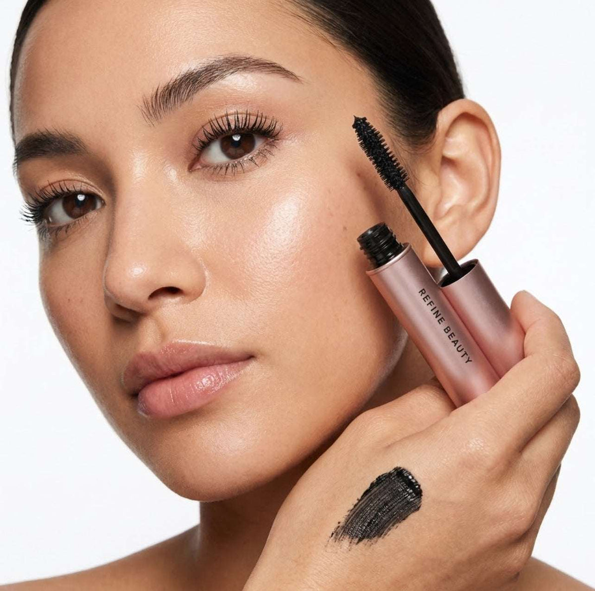 Woman applying Refine Beauty volume mascara  to her eyelashes with a close-up of the mascara tube.