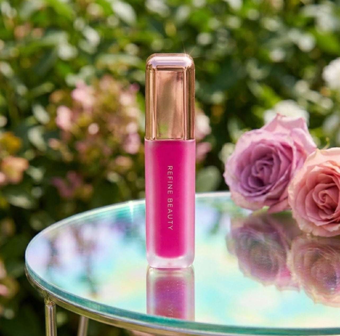 Refine Beauty blush with rose gold cap on a reflective surface surrounded by pink roses and green foliage.