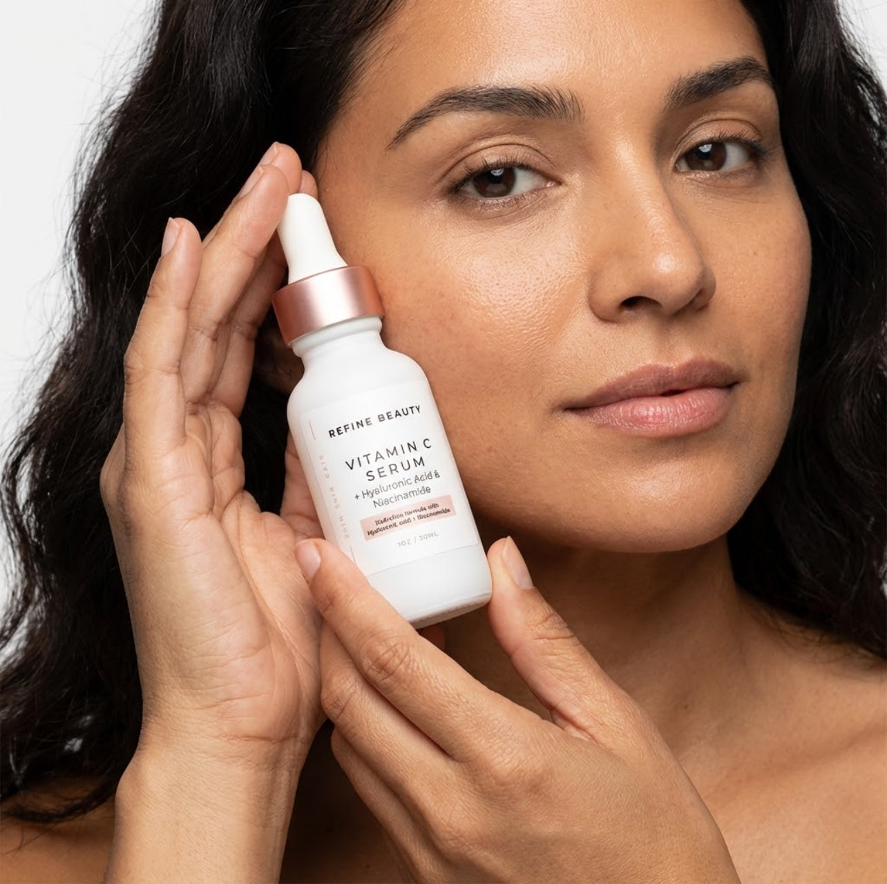 Woman holding a Refine Beauty Vitamin C Serum skincare serum bottle near her face against a neutral background
